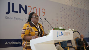 Dr. Lydia Dsane-Selby speaks at a podium on stage at the 2019 JLN Global Meeting.